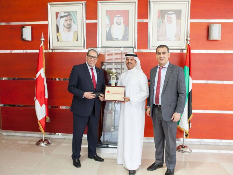 Three individuals in business attire stand together and pose, with one holding a certificate. One man is dressed in traditional UAE dress. Portraits and the Canadian and United Arab Emirates flags are displayed in the background.